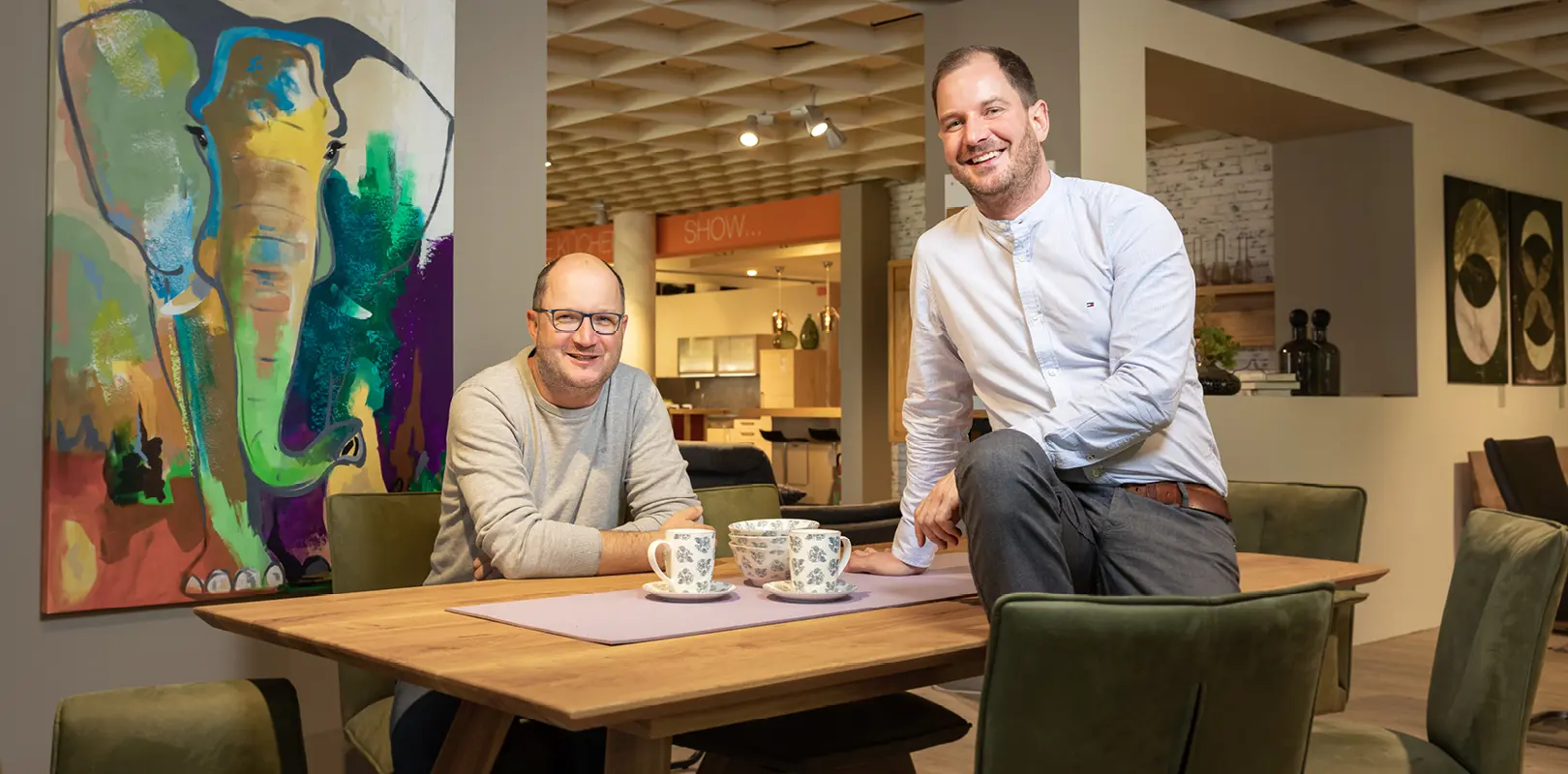 zwei freundliche Männer an Esstisch, lachend in Einrichtungshaus